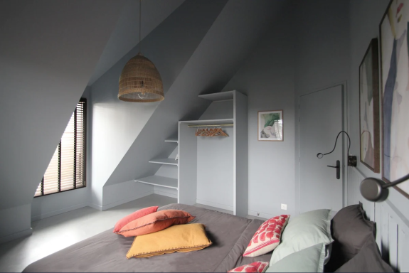 Chambre aux murs gris, lit avec coussins colorés, étagères et lumière naturelle.